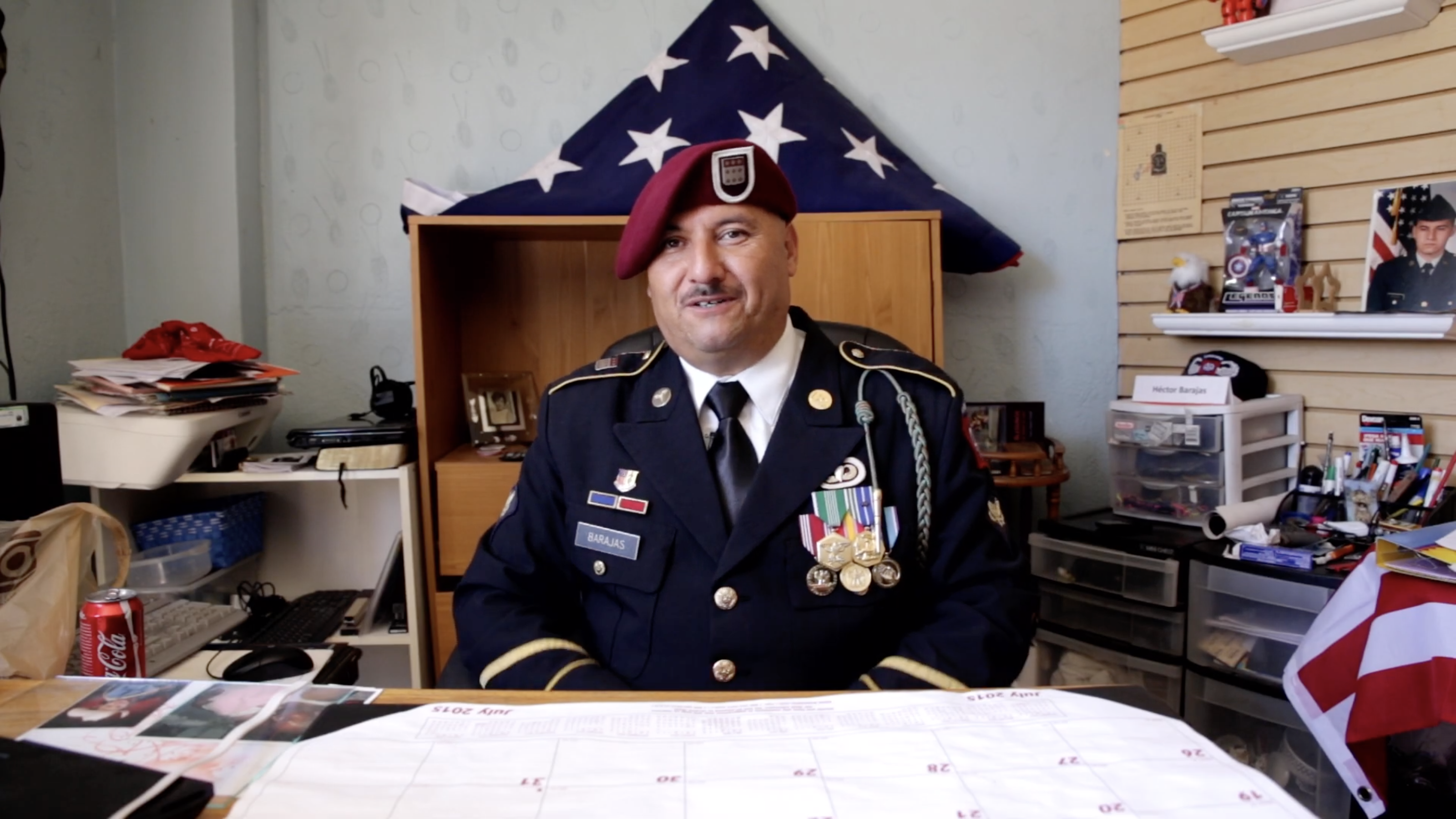 Hector Barajas wearing in his U.S. Marine uniform seated at his desk, an American flag folded into a triangle on a shelf behind him