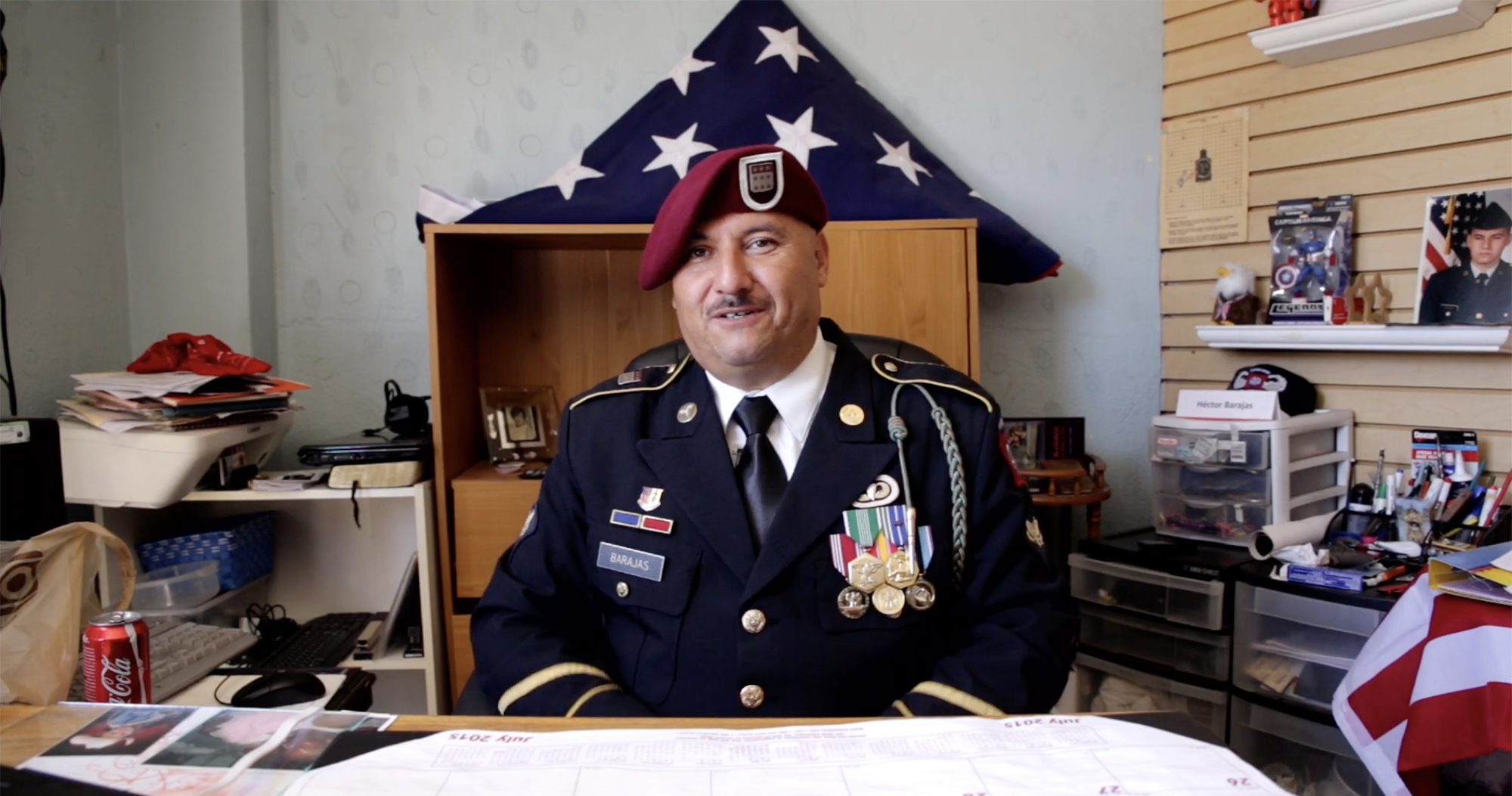 Hector Barajas wearing in his U.S. Marine uniform seated at his desk, an American flag folded into a triangle on a shelf behind him