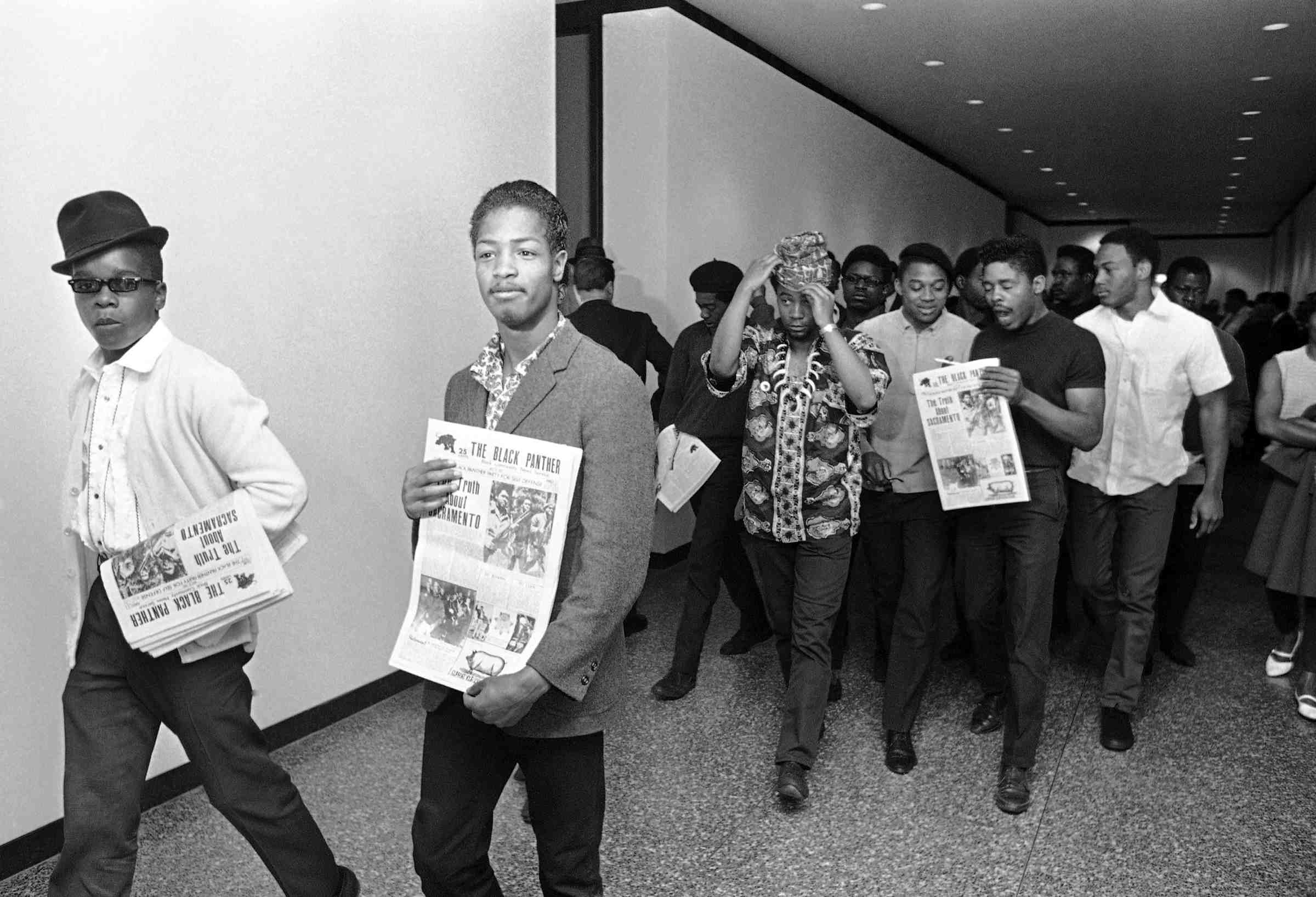 Members of the Black Panther Party walking down a hallway, holding newspapers.