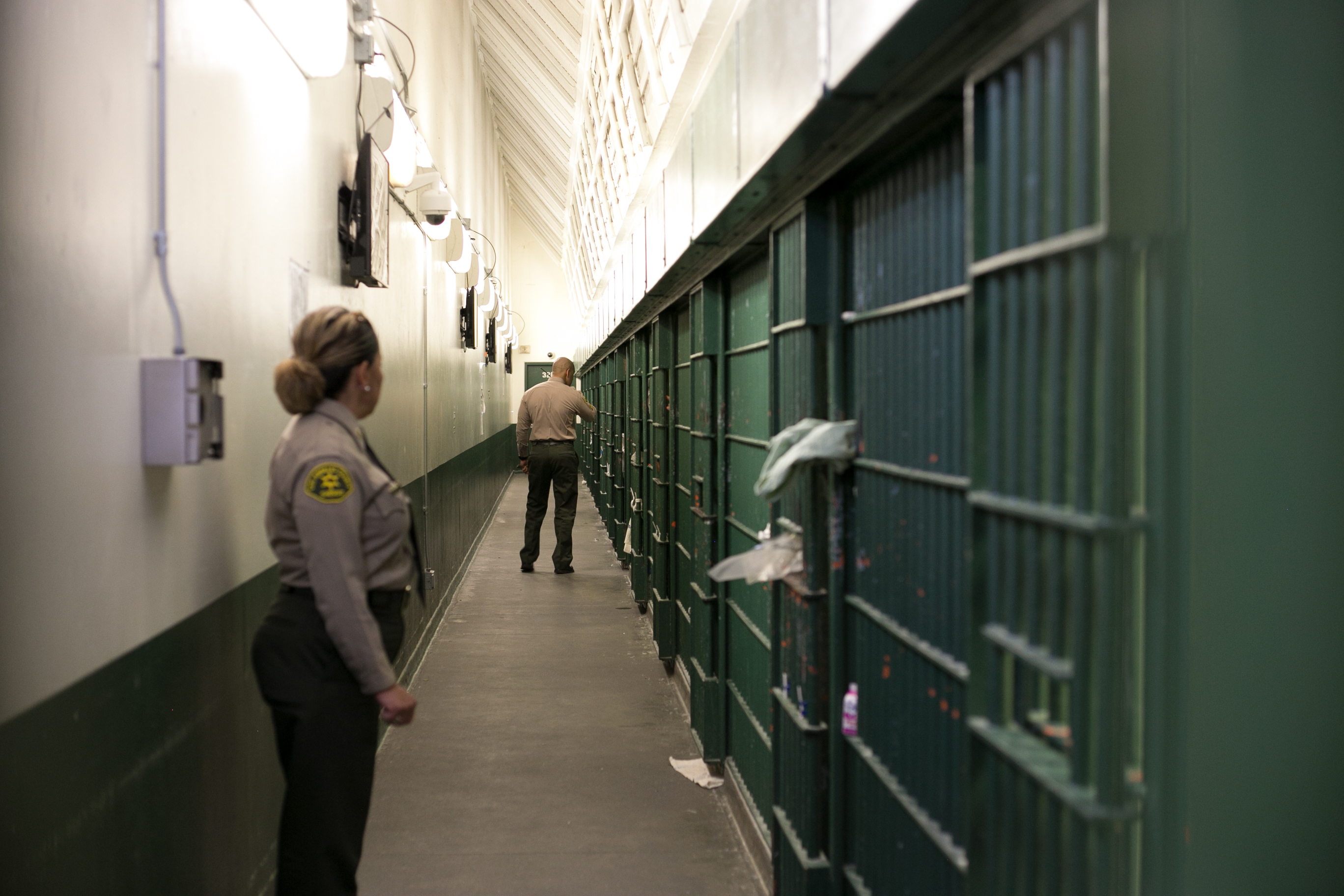 A female sheriff's deputy standing in front of a jail cell, she's looking to her left where a male sheriff's deputy is walking down the hall with his back to her.