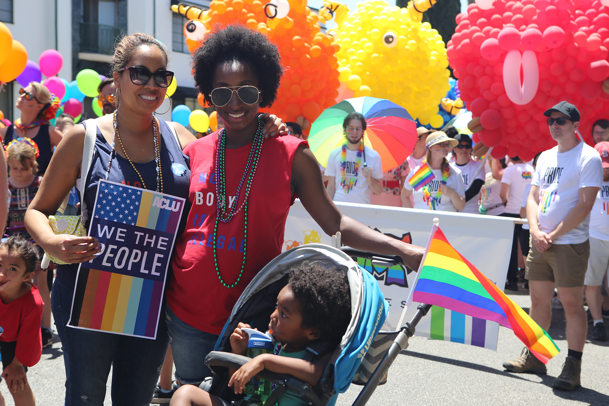 Two women with a child in a stroller in the foreground at a Pride Parade. One woman is holding a sign that reads We the People.