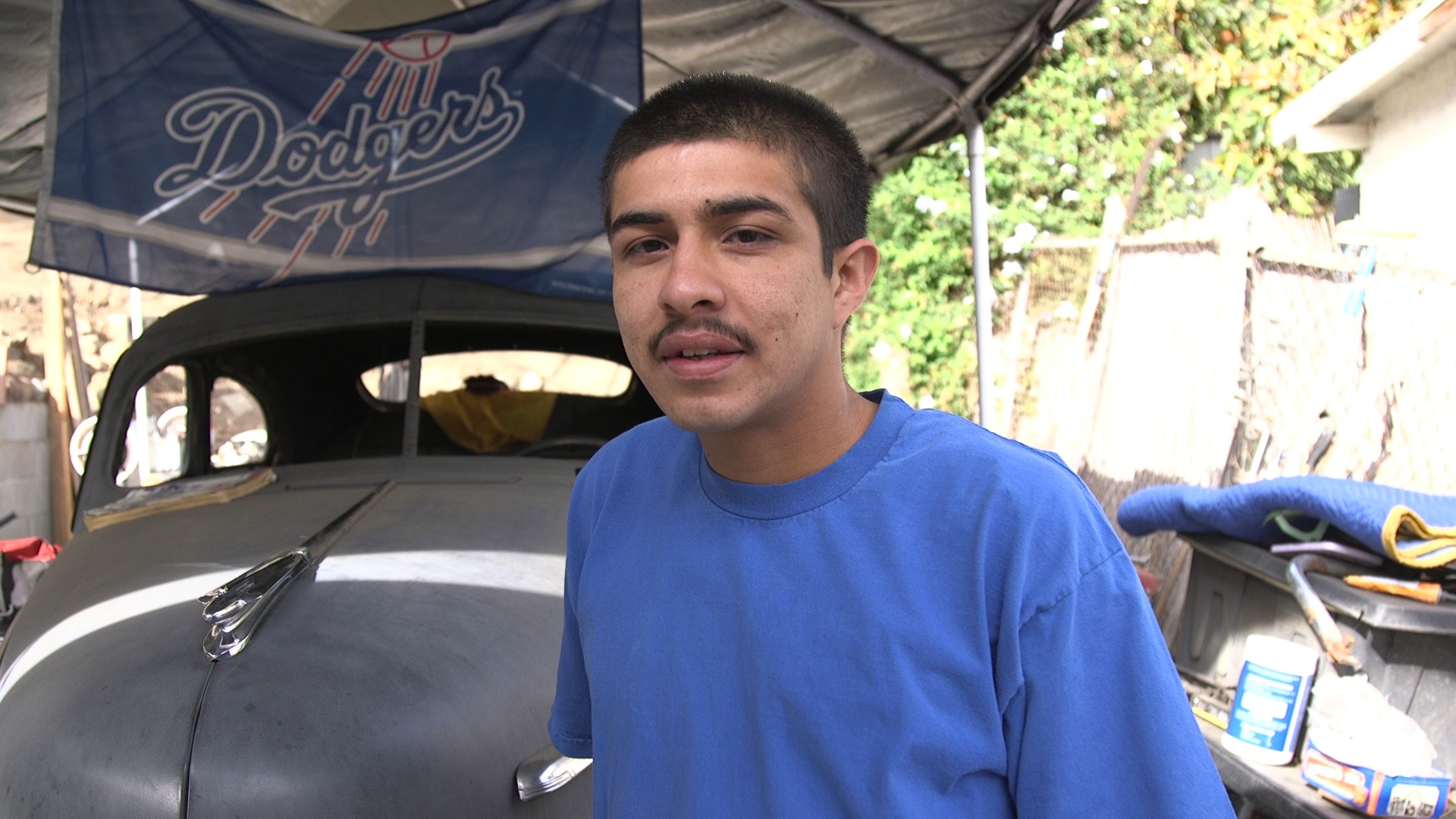 Peter Arrellano, plaintiff in YJC v. Los Angeles, standing in front of a Volkswagen bug in a carport with a Dodgers flag hanging from the top of the carport