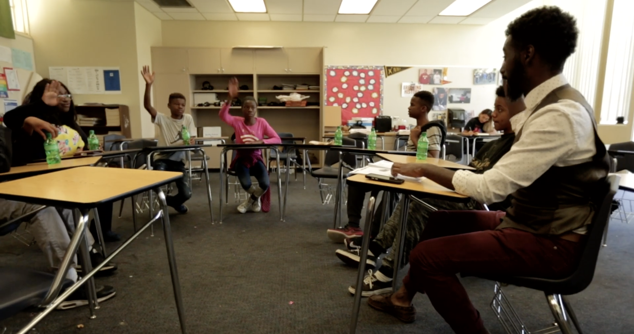 Six youth and an adult sitting in a classroom with their desks forming a semi-circle. Three of the youth have their hands raised. 