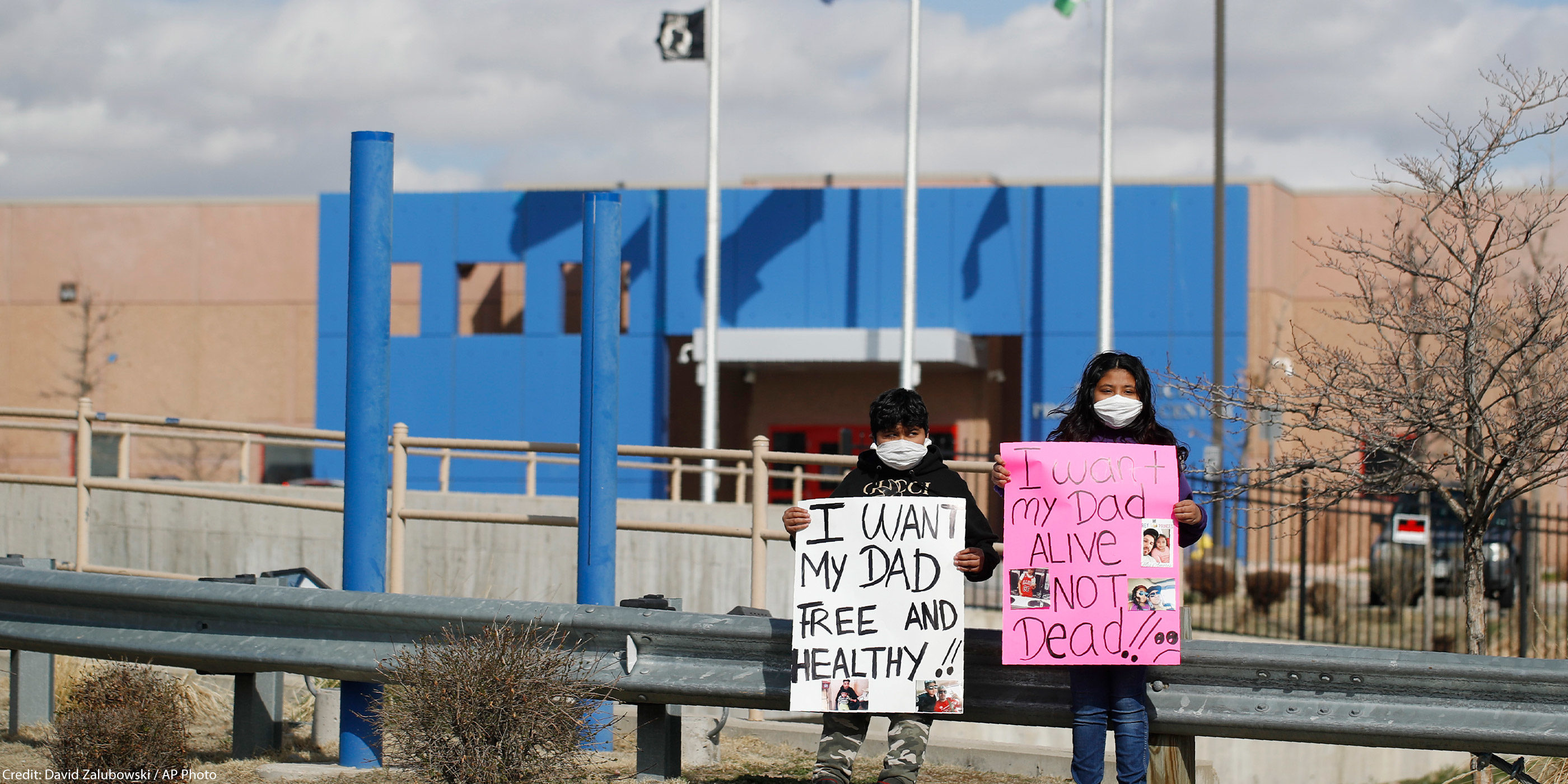 Two children wearing face masks standing in front of an immigration detention center holding signs that read: "I want my dad free and healthy" and "I want my dad alive not dead."