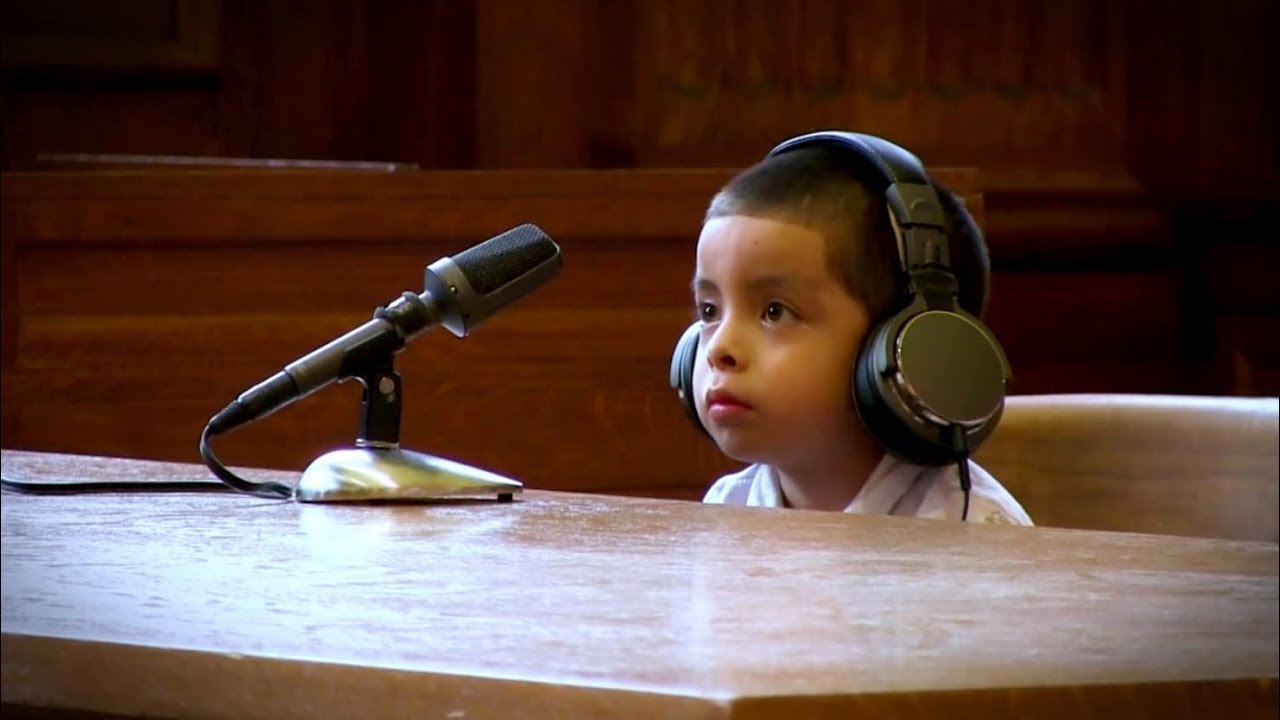 Young child in court wearing headphones, sitting at a table with a microphone in front of him.