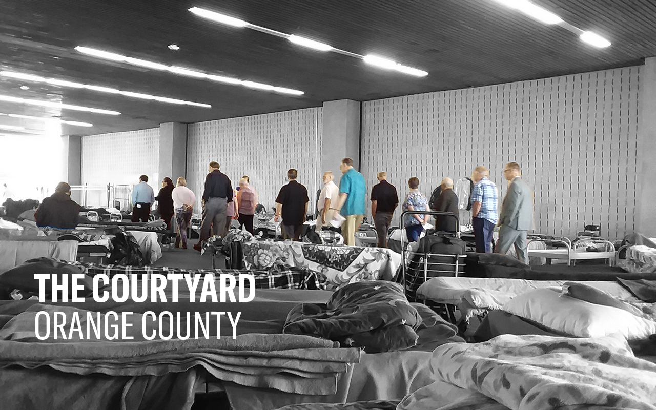 A scene from a homeless shelter in Orange County. In the foreground, cots with blankets and pillows a couple feet apart. In the background, a group of people standing in a line.