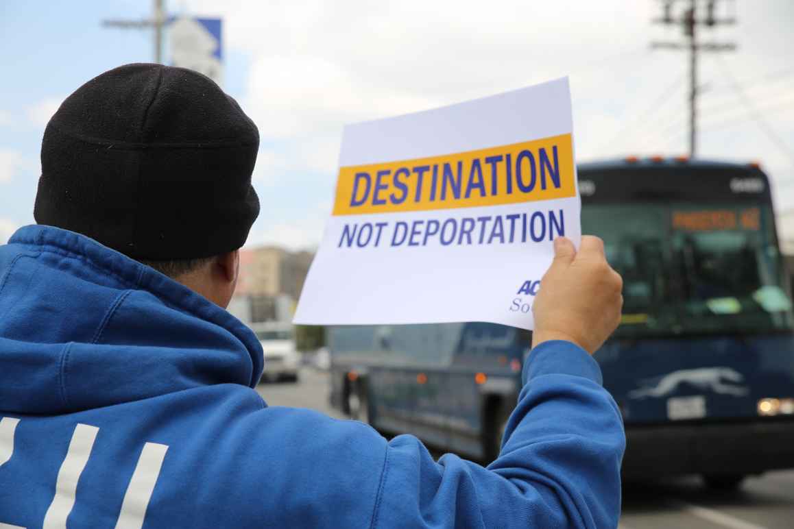 Man on sidewalk, holding a sign as a  Greyhound bus passes by: "Destination not deportation"