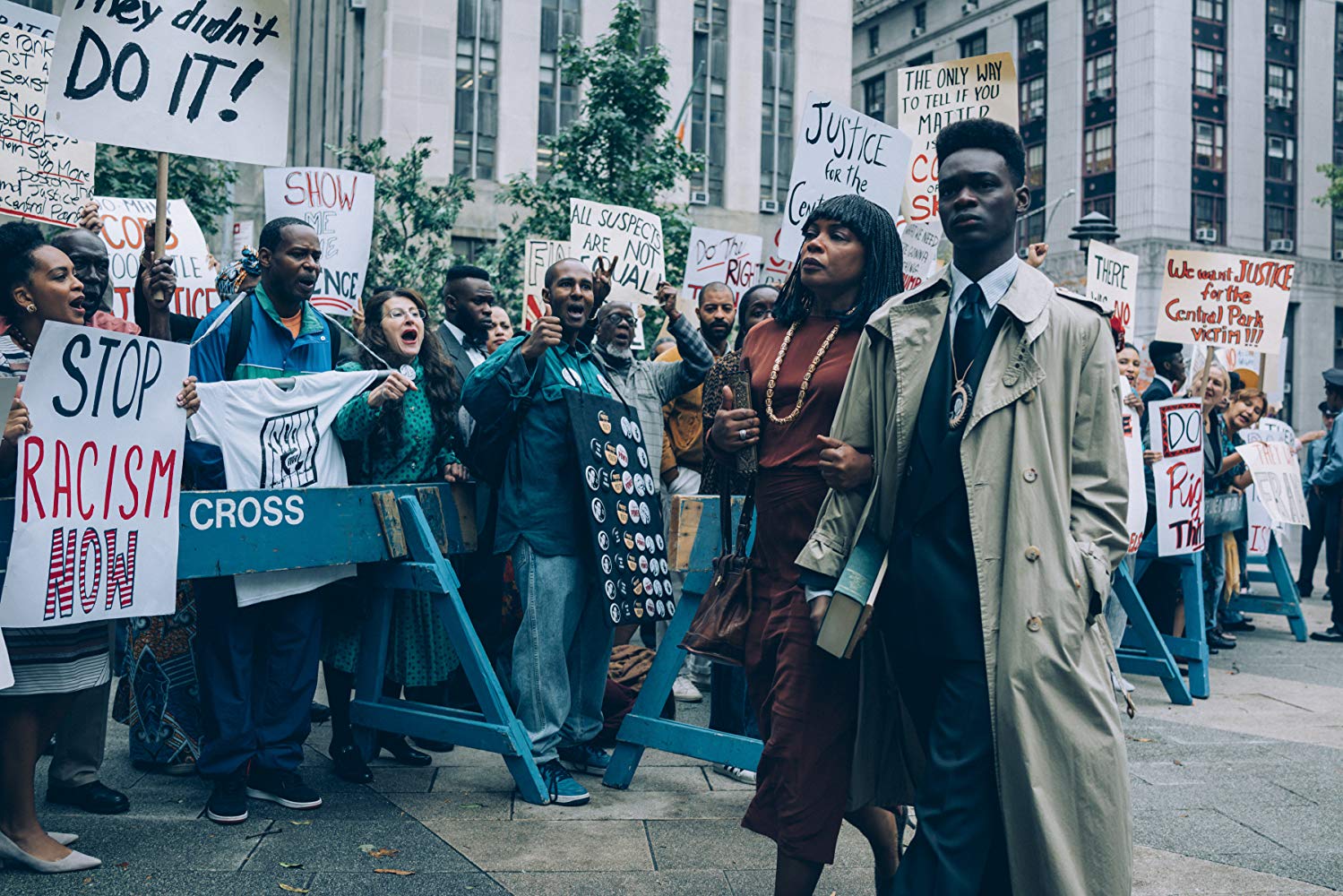 A production still of the Netflix series "When They See Us": A Black man and woman walk arm in arm alongside protestors behind a barricade holding posters that read "They didn't do it" and "Stop racism now."